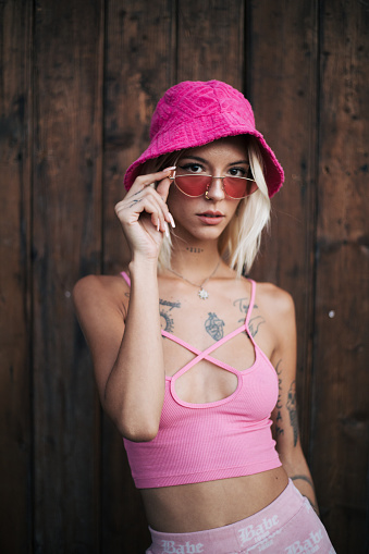 Blonde woman posing with tattoos all over her body in front of a wooden wall. Colourful and styled pink outfit.