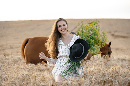 A young girl enjoys a beautiful summer day in early August and walks her cool horse in a golden field collecting flowers for a bouquet