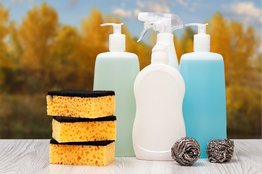 Plastic bottles of dishwashing liquid, tile cleaner, detergent for microwave ovens and stoves, sponges in front of the autumn background. Washing and cleaning concept.