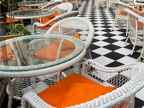 Close-up image of rows of glass topped garden patio table with rattan wickerwork chairs outdoors, restaurant facade, al fresco dining area, black and white checked tiled floor, focus on foreground