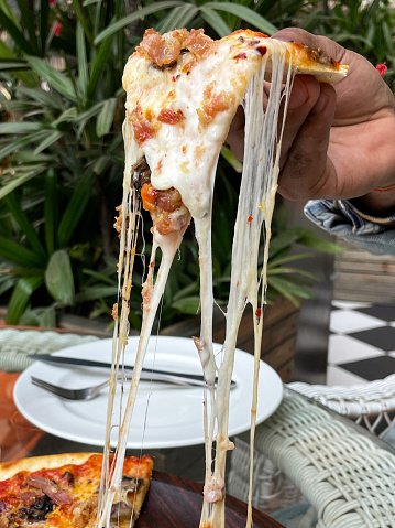 Stock photo showing a close-up view of unrecognisable person holding a triangular slice of pizza with stringy melted buffalo mozzarella in restaurant.