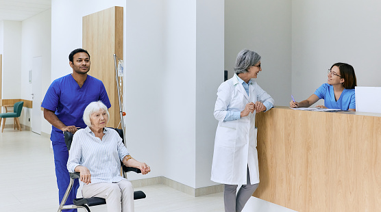 Healthcare assistant pushes wheelchair with senior woman in motion along corridor of hospital near reception desk with on-call nurse and doctor. Patient care in hospital