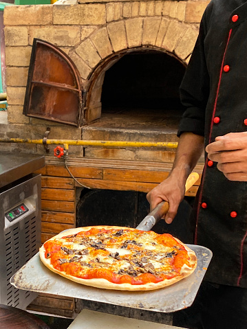 Stock photo showing close-up view of freshly cooked pizza base covered with rich tomato sauce, melted buffalo mozzarella cheese and mushroom slices being removed from traditional wood burning brick pizza oven on metal pizza paddle shovel.