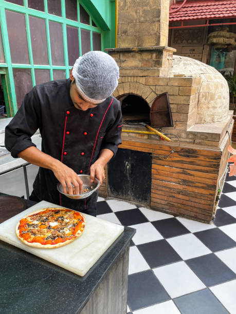 Stock photo showing close-up, elevated view of freshly cooked pizza base covered with rich tomato sauce, melted buffalo mozzarella cheese and mushroom slices  having a topping of chopped ham added by hand of unrecognisable chef.