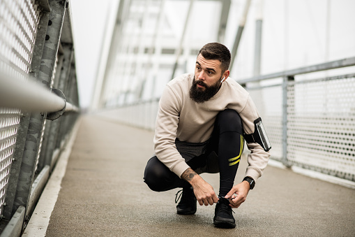 Young man with a beard on his morning run on a bridge over a river, using modern technology. Handsome young runner using phone holder on his arm, and Air pods for his morning run on a rainy day.