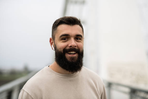 Young man with a beard on his morning run on a bridge over a river, using modern technology. Handsome young runner using phone holder on his arm, and Air pods for his morning run on a rainy day.