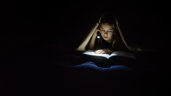 A girl with a flashlight reads a book under a blanket in the dark.