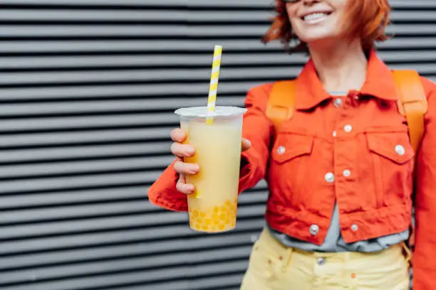 Glass of fruity sugar flavored tapioca pearl bubble tea with straw in hand of no face hipster fashion woman in bright clothes on the gray striped wall background. Selective focus. Copy space