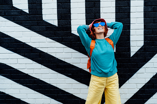 Happy emotional hipster fashion young woman in bright clothes, heart shape sun glasses and bucket hat posing on the painted brick wall background. Urban city street fashion. Selective focus