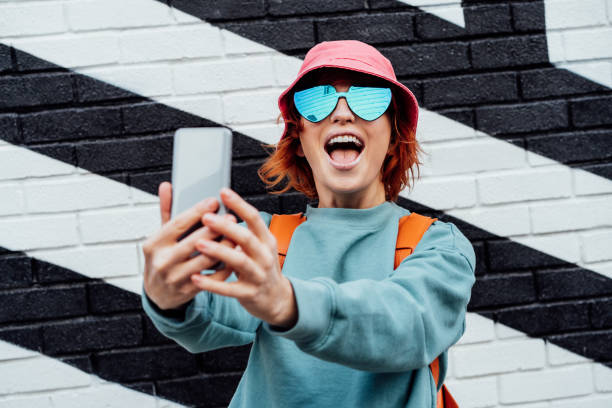 Excited redhead woman screaming while taking a selfie photo outdoors. Emotional hipster fashion women in bright clothes, heart shaped glasses, bucket hat taking selfie photo on the phone camera Excited redhead woman screaming while taking a selfie photo outdoors. Emotional hipster fashion women in bright clothes, heart shaped glasses, bucket hat taking selfie photo on the phone camera. bucket hat stock pictures, royalty-free photos & images