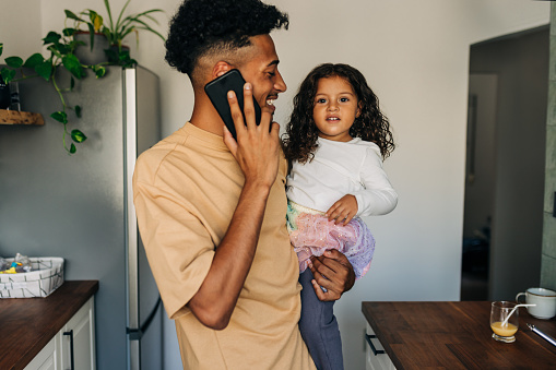 Happy young dad smiling and speaking on the phone while carrying his daughter. Cheerful single father spending his time at home with his young daughter.