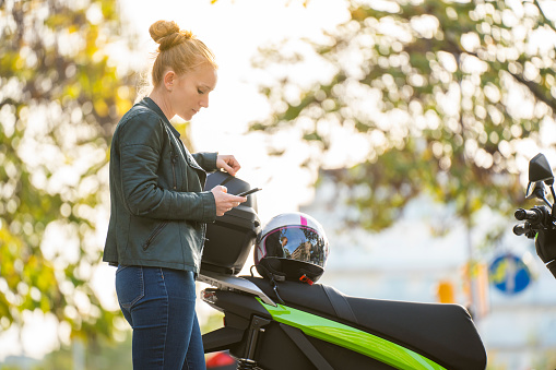 Portrait of red-haired woman using the smartphone next to her parked motorcycle