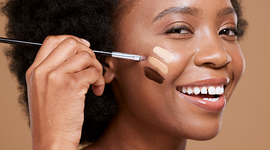 Black woman, face and smile for makeup foundation, cosmetics or skincare against a brown studio background. Portrait of happy African female applying cosmetic beauty product with brush for skin toner