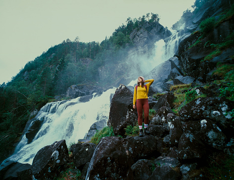 Young Caucasian woman  standing near the powerful  waterfall in mountains during her hike in Norway