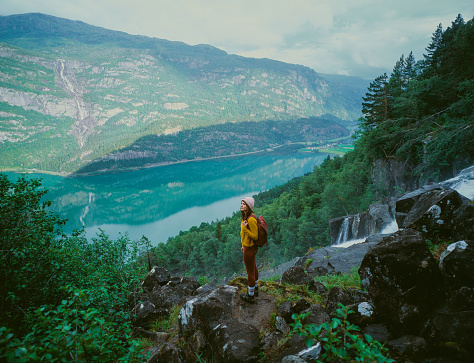 Young Caucasian woman  standing near the powerful  waterfall in mountains during her hike in Norway