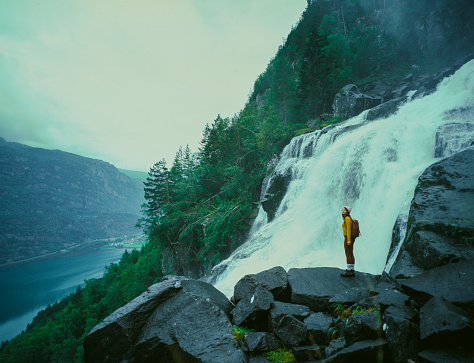 Young Caucasian woman  standing near the powerful  waterfall in mountains during her hike in Norway