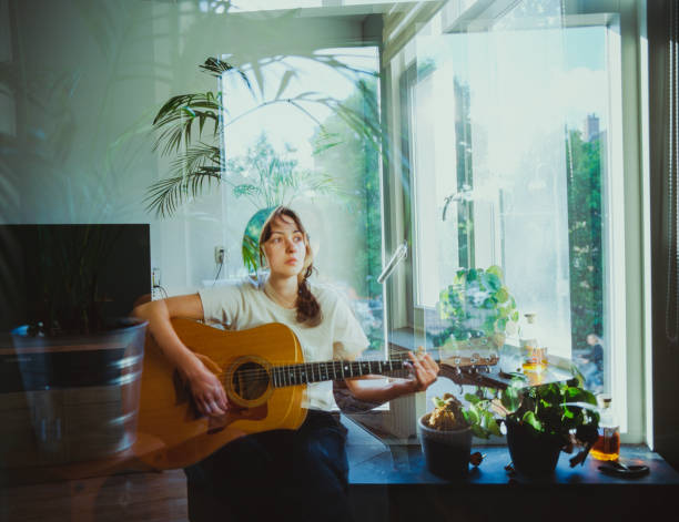Young Caucasian woman sitting on windowsill and playing guitar