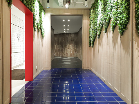Hallway of a building with blue tiles and wooden texture walls. Artificial greenery on the walls.