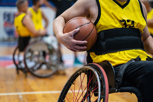 An unrecognizable basketball player using a wheelchair with a basketball under his arm, more team members in the background