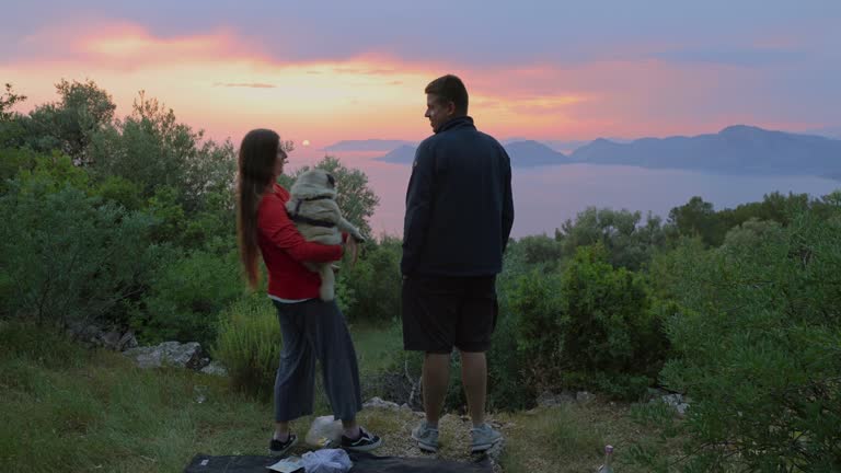 A woman and man with a dog admiring sunset overlooking the sea with mountains during sunset in Turkey