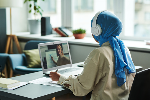 Back view of young Muslim female student in headphones and hijab listening to explanation of teacher during online lesson