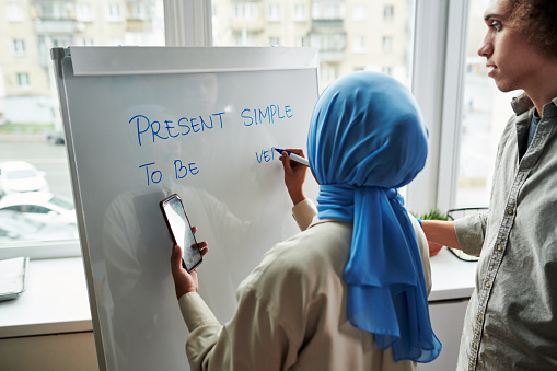 Back view of young Muslim female teacher explaining multi-ethnic guy Present Simple tense while wiriting down examples on whiteboard