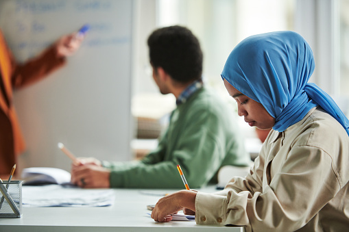 Side view of young serious Muslim woman in blue hijab making notes in copybook while sitting by workplace during presentation