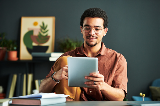 Young Middle Eastern male student looking through online information on screen of tablet while sitting by workplace with books