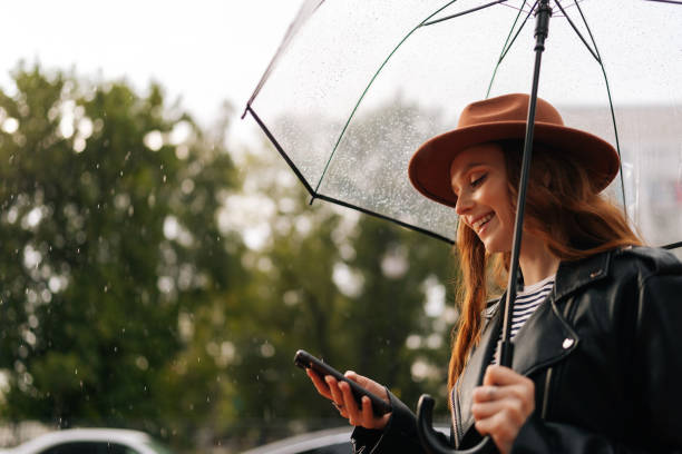 istock Side view of cheerful young woman wearing elegant hat using typing smartphone smiling looking away standing with transparent umbrella. 1443306023