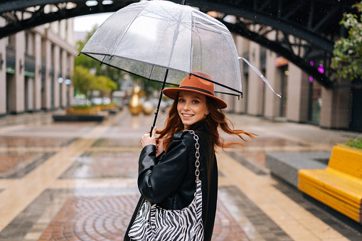View from shoulder to positive elegant woman in fashion hat walking on beautiful city street with transparent umbrella enjoying rainy weather outdoors, looking at camera. Concept of female lifestyle.