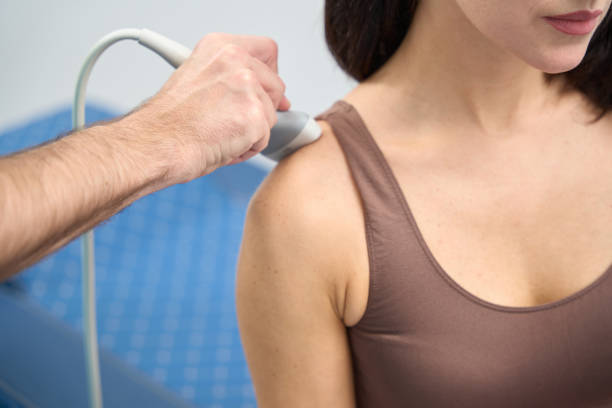 Woman is undergoing shoulder examination on clinic with ultrasound machine Calm female patient sitting on a medical examination while the doctor runs a medical device over the upper part of the shoulder shoulder scan stock pictures, royalty-free photos & images