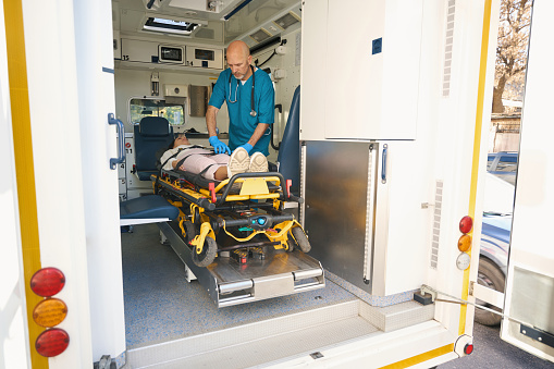 Focused man preparing woman for transportation for medical care while she lies on stretcher in car interior