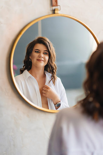 a young brunette woman in a white shirt looks at her reflection in a round mirror. care and decorative cosmetics for women. cosmetologist services. furniture and decor for the house.
