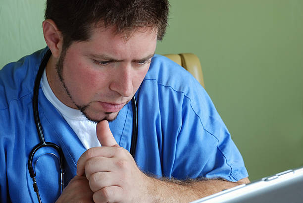 Doctor with stethoscope looking at computer #1 Doctor with stethoscope and blue scrubs intently looking at computer medical scribe remote stock pictures, royalty-free photos & images