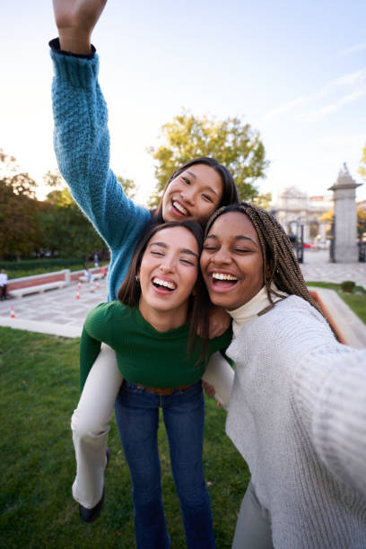 Happy photo of three diverse female friends taking cheerful selfie doing piggyback. Girls having fun Vertical photo of three multicultural female friends taking smiley selfie doing piggyback. Young women having fun together outdoors at city park in Madrid. People enjoying travel in weekend holidays. selfie madrid photos stock pictures, royalty-free photos & images