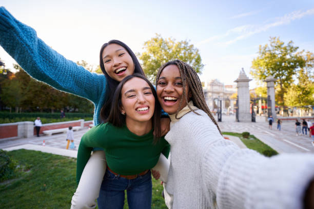 Happy photo of three diverse female friends taking cheerful selfie doing piggyback. Girls having fun Playful photo of three multicultural female friends taking smiley selfie doing piggyback. Young women having fun together outdoors at city park in Madrid. People enjoying travel in weekend holidays. selfie madrid photos stock pictures, royalty-free photos & images