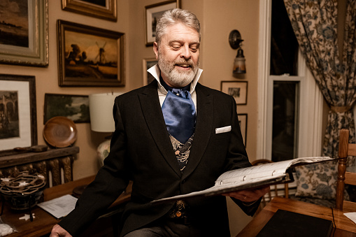 Mature man practising singing for Christmas carolling.  He is dressed up in period costume. Interior of vintage home during evening.