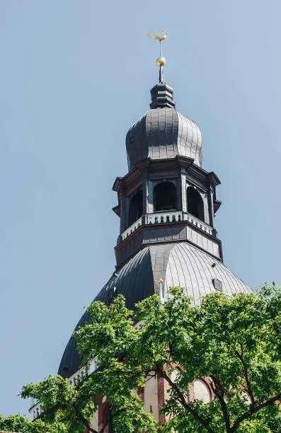 Photo of church tower with a green metal roof and a balcony topped with a gilded rooster under a blue sky.