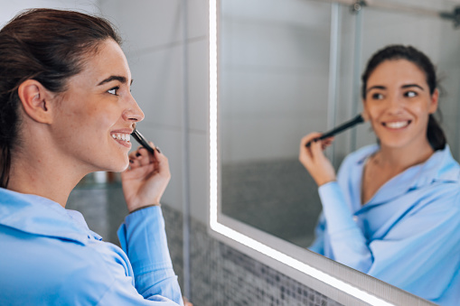 A young Caucasian woman is standing in her bathroom and cheerfully applying make-up while looking at her reflection in the mirror.