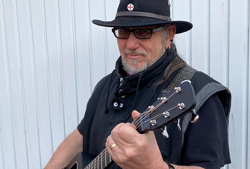 Older man dressed in black plays a black guitar. He wears a black cowboy hat with a Templar pin