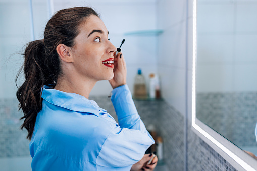 A young Caucasian woman is looking at the mirror and applying mascara, while standing in her bathroom.