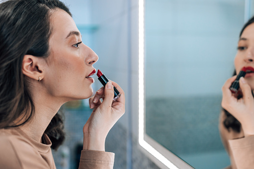A young Caucasian woman is applying red lipstick in the bathroom mirror with a focused facial expression.