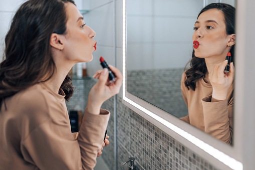 A young Caucasian woman is looking at her mirror reflection and puckering her lips while applying make-up in her bathroom.