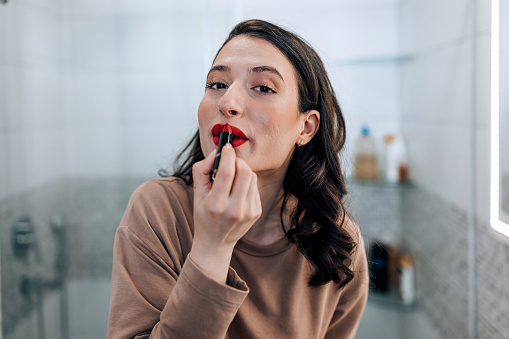 A young Caucasian woman is standing in her bathroom and looking at the camera while carefully applying lipstick.