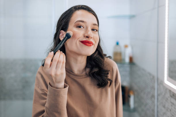 Love how this look turned out A beautiful, young Caucasian woman ith dark hair is looking at the camera with a smirk while applying blush. rosy cheeks stock pictures, royalty-free photos & images