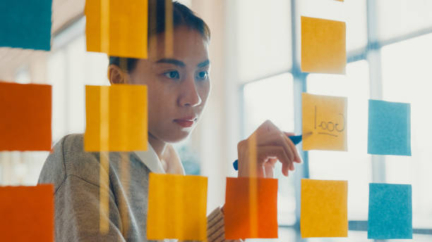 Asian girl focus on transparent board writing roadmap planning on sticky note work outside office in co-working room. Asian girl focus on transparent board writing roadmap planning on sticky note work outside office in co-working room. Business Woman happy working at co-working space. Alternatives workspace concept. mirror writing stock pictures, royalty-free photos & images
