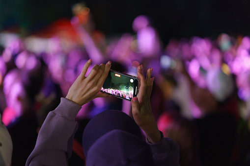 People are excited during street rock concert. They dance. Bright blue neon lighting. Night life of the big city. Young woman makes photos via smartphone. Festive mood. Selective focus. Back view