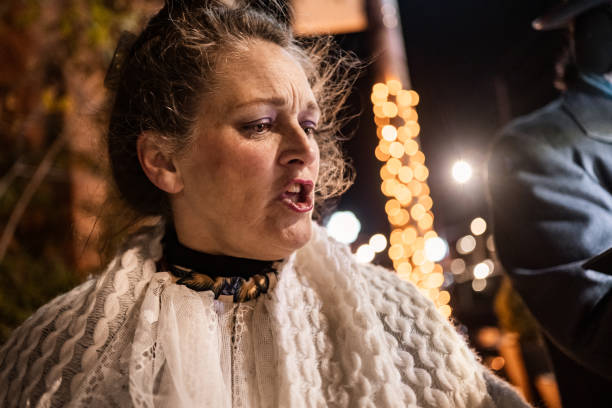 Mature people singing Christmas carols outdoors at night.  They are dressed up in period costumes. Exterior of city street with Christmas decor.