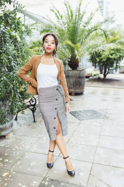 Young African American mixed race woman in brown jacket, midi pencil skirt with front slit, high heel shoe with belt strap in park or street outdoor. White t shirt mock up. Full body fall portrait