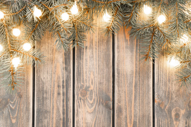 istock Burning lit light bulbs of Christmas tree branches garland lying on plank surface. Rustic holiday background, copy space 1443337629